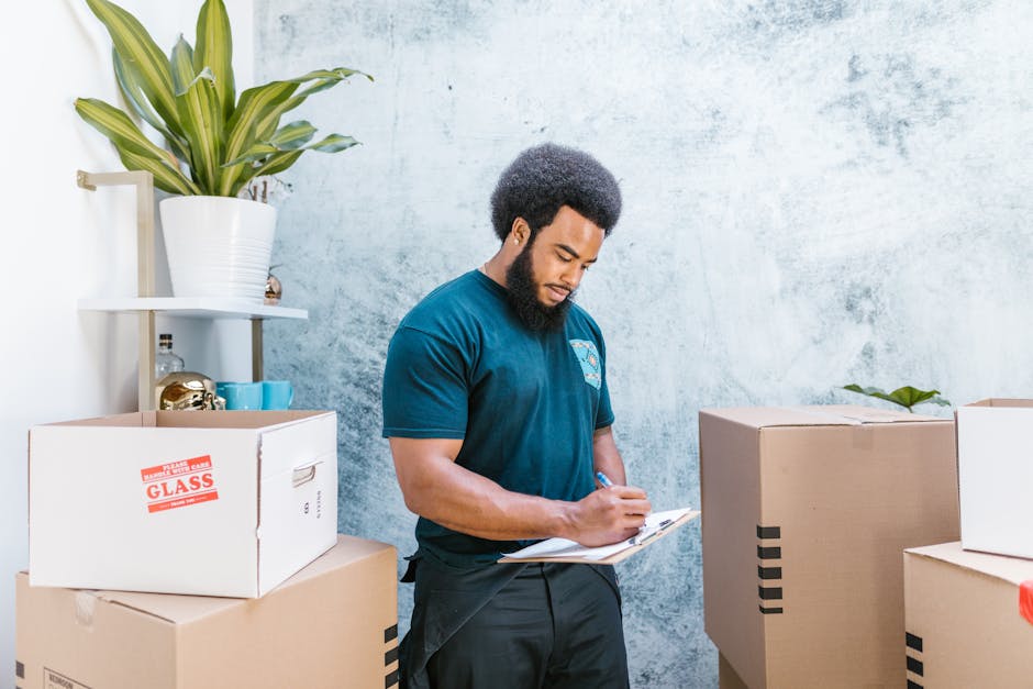 A collection of packed moving boxes and packaging materials placed on a wooden floor inside a property, ready for home relocation. The boxes are mostly cardboard, some with handwritten labels such as