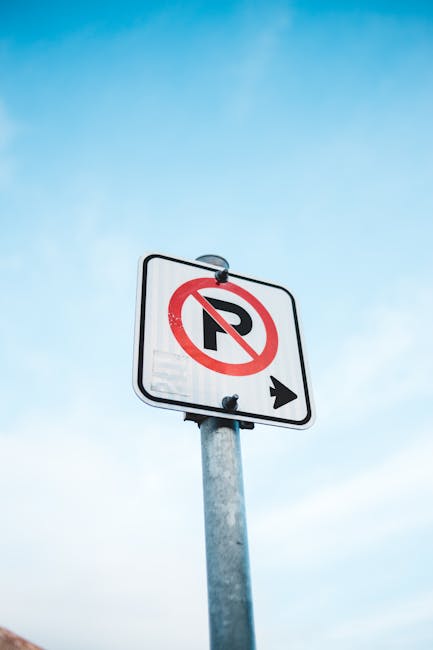 A rectangular 'No Parking' sign with a white background, red border, and black text is mounted on a metal pole situated outdoors. Below the text, two black arrows point left and right, indicating park