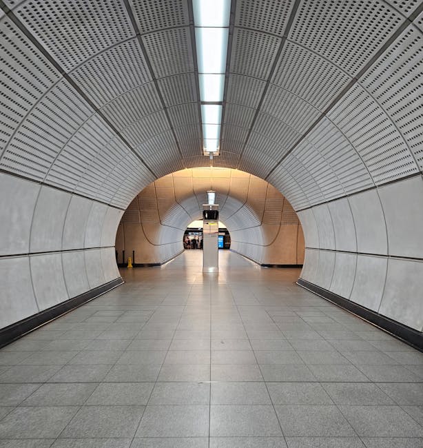 A nighttime scene at a train station platform showing a row of parked trains with blue and white carriages on the left side. The platform has a textured yellow tactile paving strip along the edge for